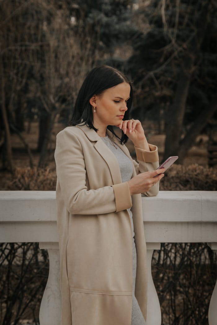 Side view of peaceful female in coat using mobile phone while standing in park in fall on cloudy day